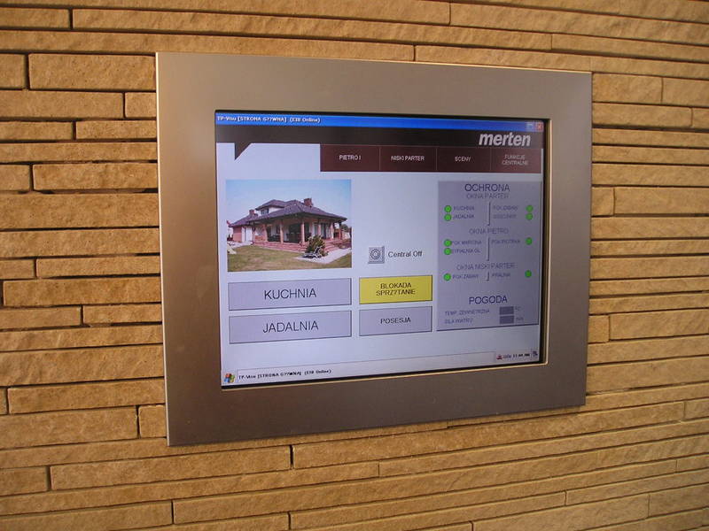 Panel dotykowy systemu inteligentnego domu Merten zamontowany na ścianie z dekoracyjnej cegły, wyświetlający schemat domu, opcje sterowania oświetleniem, ochroną i pogodą.