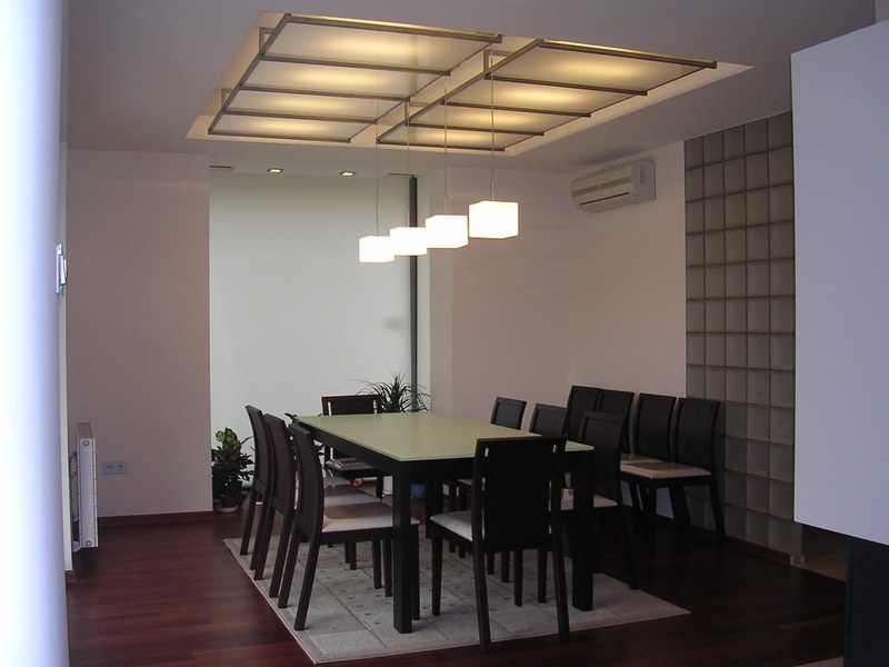 Nowoczesna jadalnia z podwieszanym sufitem z oświetleniem rastrowym i wiszącymi lampami kostkami, ściana z luksferów, parkiet z ciemnego drewna.