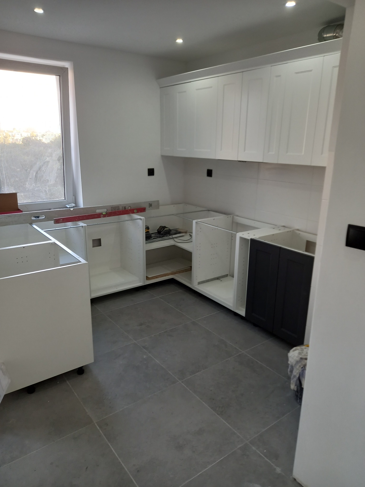 Montaż białych i grafitowych szafek kuchennych w Krusze, widoczne narzędzia i poziomica, płytki podłogowe w odcieniach szarości, okno z widokiem na zewnątrz.