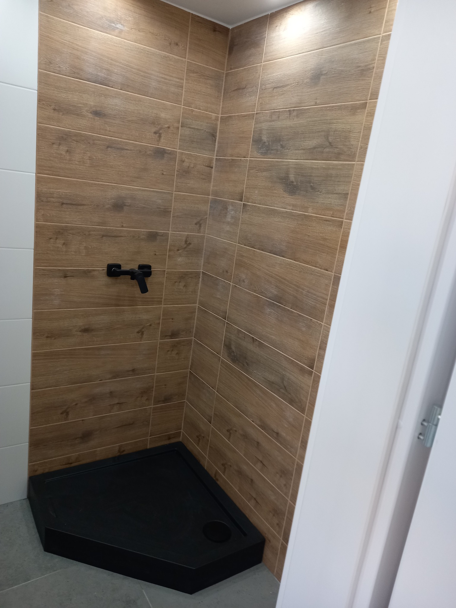 Wykończona kabina prysznicowa z czarnym brodzikiem i baterią, ściany wyłożone płytkami imitującymi drewno, kontrastujące z białymi płytkami i ościeżnicą.