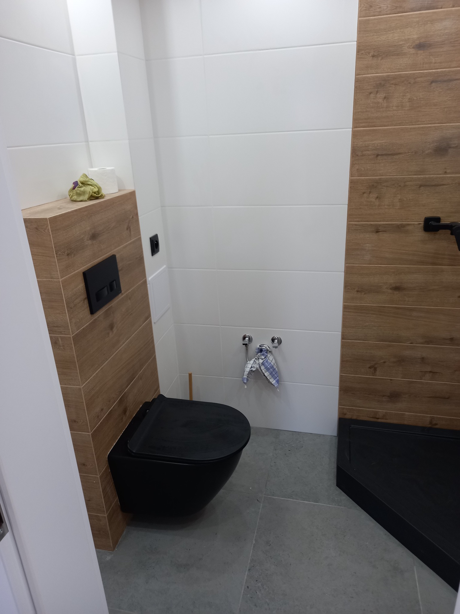 Nowoczesna łazienka z czarną toaletą wiszącą i kabiną prysznicową, wykończona białymi i drewnopodobnymi płytkami, szara podłoga.