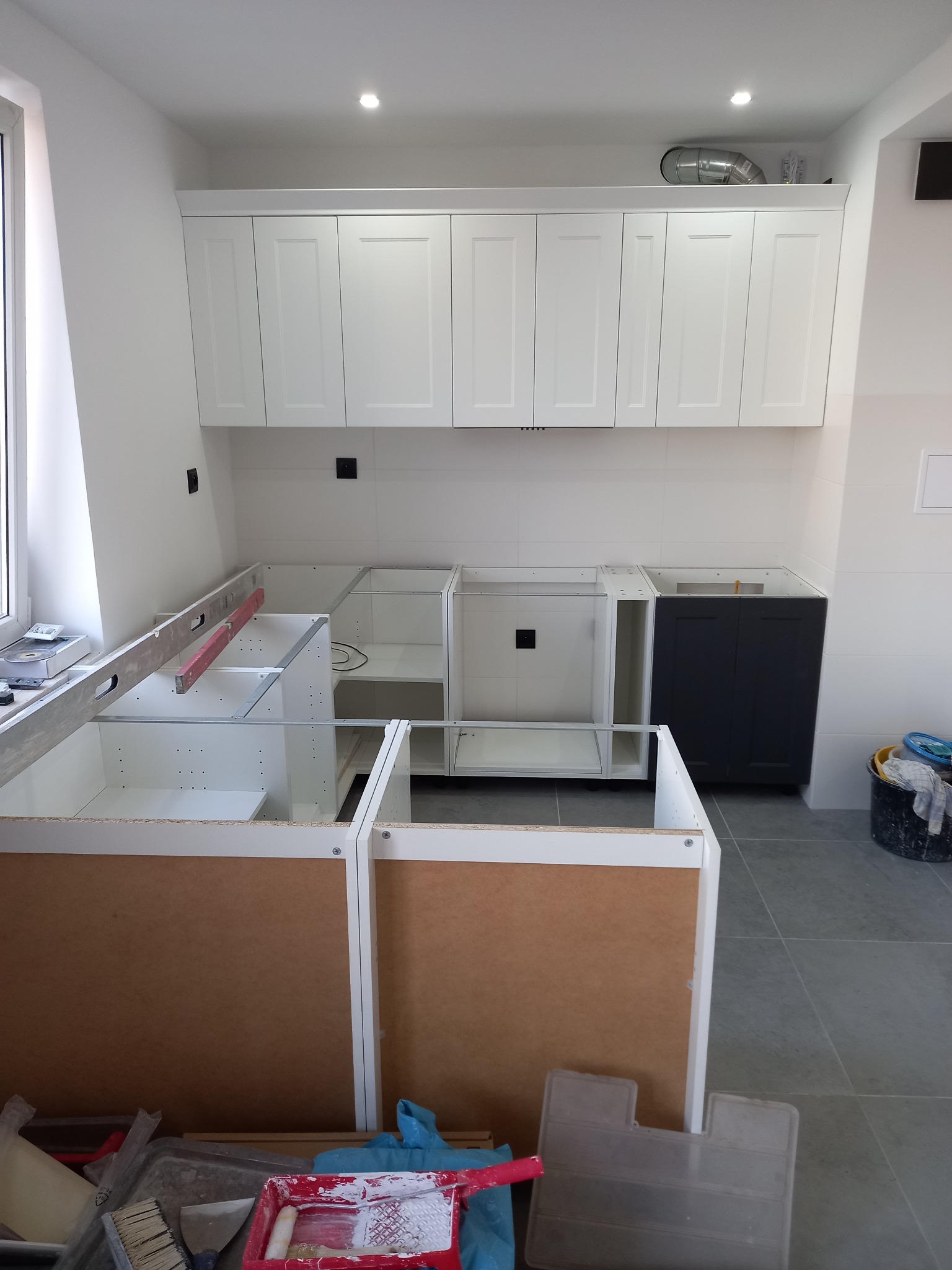 Montaż szafek kuchennych: białe szafki wiszące i dolne w trakcie instalacji, poziomica na blacie, widoczne narzędzia malarskie i wiadro w tle.
