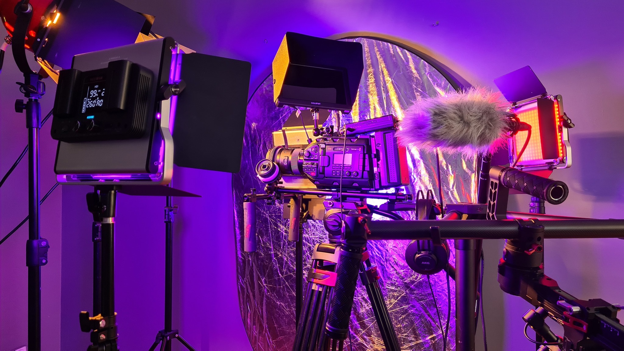 Profesjonalny sprzęt filmowy: kamera z mikrofonem typu 'deadcat', monitor podglądowy i oświetlenie LED ustawione na statywach, tło z folii odblaskowej, fioletowe oświetlenie sceniczne.
