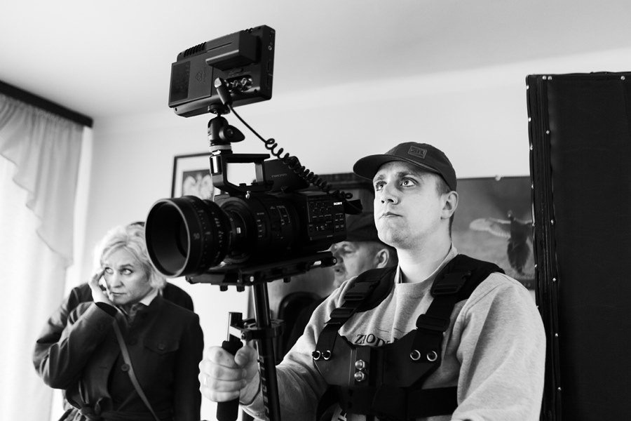 Czarno-białe zdjęcie kamerzysty z profesjonalną kamerą Sony na statywie, w tle kobieta rozmawiająca przez telefon i fragment scenografii.