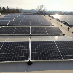 Rząd paneli słonecznych na płaskim dachu budynku, z widocznymi mocowaniami, okablowaniem i krajobrazem w tle, oświetlonych popołudniowym słońcem.