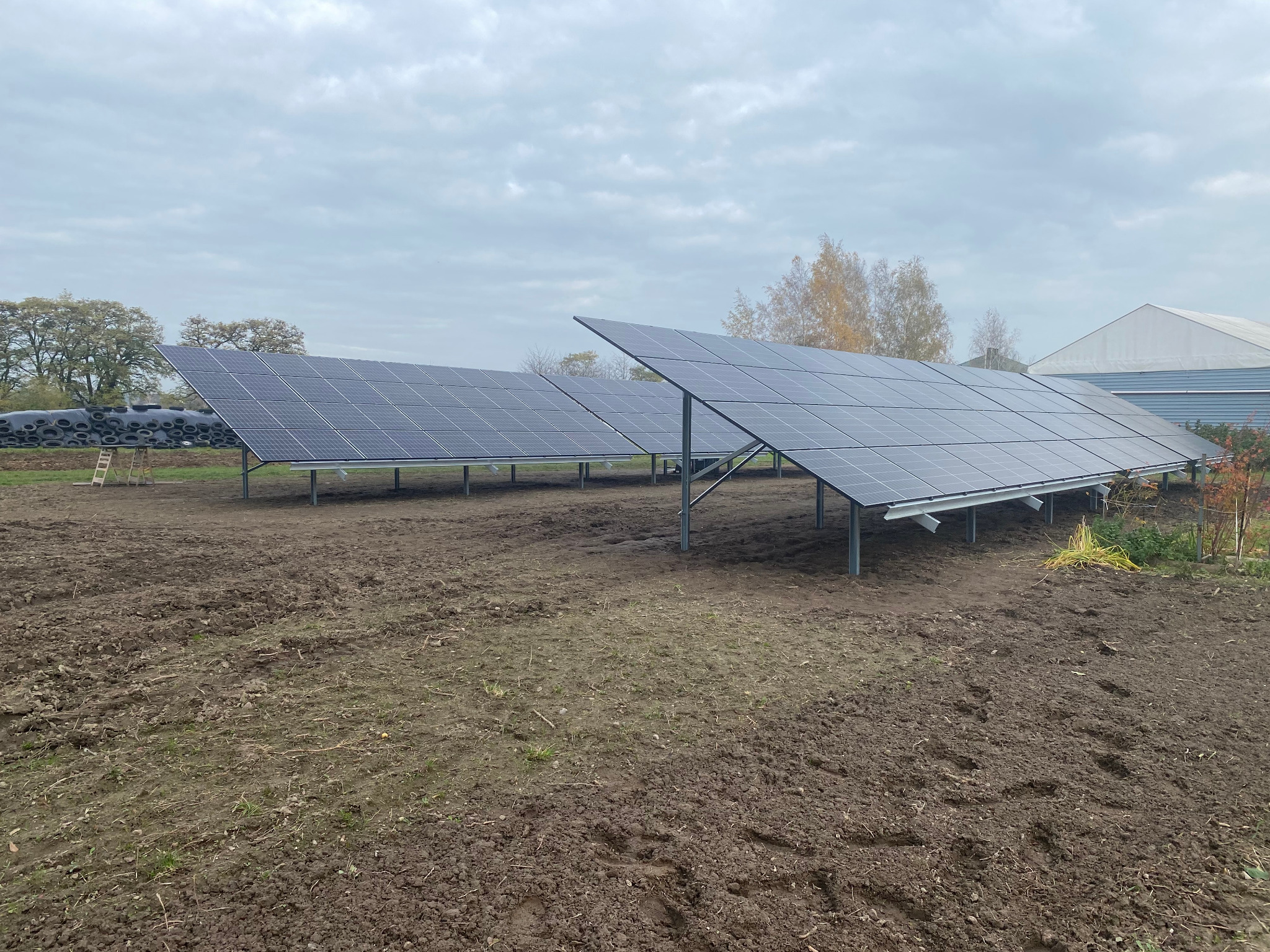 Dwa rzędy paneli fotowoltaicznych na polnej działce z widocznymi śladami prac ziemnych i stertą opon w tle, pochmurne niebo.