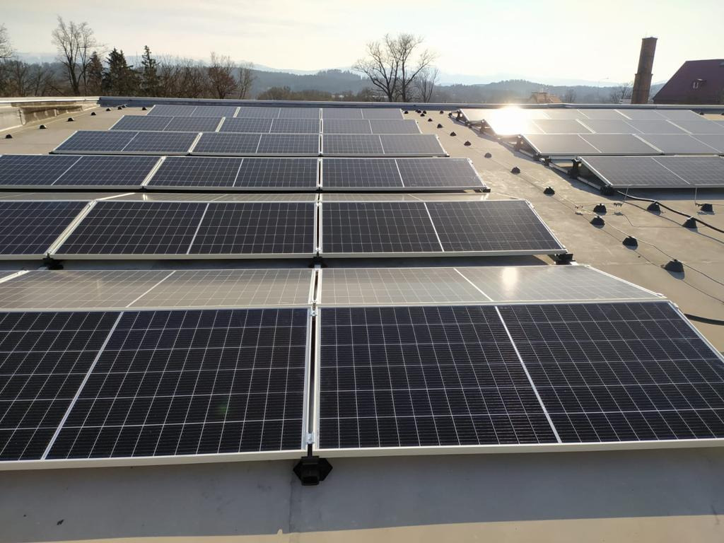 Rząd paneli słonecznych na płaskim dachu budynku, z widocznymi mocowaniami, okablowaniem i krajobrazem w tle, oświetlonych popołudniowym słońcem.