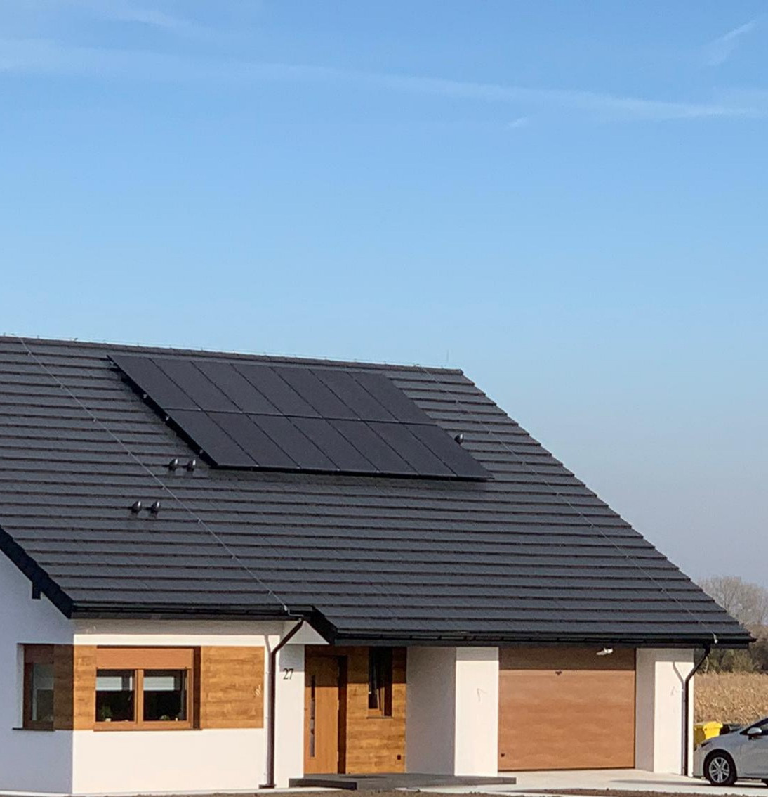 Czarny panel słoneczny na grafitowym dachu nowoczesnego domu jednorodzinnego z drewnianymi akcentami, na tle błękitnego nieba.