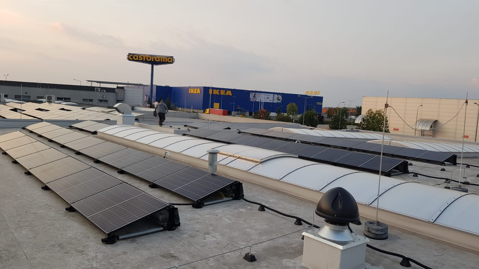 Rząd paneli słonecznych na dachu budynku z widocznymi sklepami Castorama i IKEA w tle, osoba idąca po dachu w oddali.