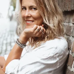 Uśmiechnięta blondynka w białej koszuli, oparta o ceglaną ścianę, z biżuterią na rękach i palcach.