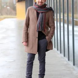 Uśmiechnięty mężczyzna w brązowym płaszczu, szarych spodniach i kolorowym szaliku stoi na zewnątrz budynku z przeszkloną fasadą, pozując z ręką w kieszeni.