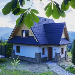 Nowo wybudowany, parterowy dom z ciemnym dachem i jasną elewacją, otoczony zielenią i górami w tle, widziany spod gałęzi drzewa.