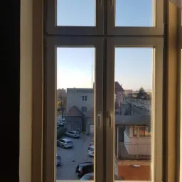 Widok z okna w Kłodzku: białe, czteroskrzydłowe okno z widokiem na parking i budynki w słoneczny dzień.