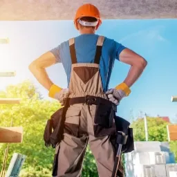 Pracownik budowlany w kasku i kombinezonie, stojący tyłem z rękami na biodrach, na tle konstrukcji dachu i błękitnego nieba.