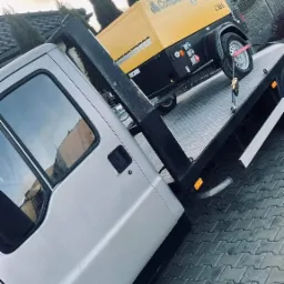 Srebrny samochód z platformą transportową przewozi żółtą sprężarkę CompAir KF-287 na tle domu z tujami.