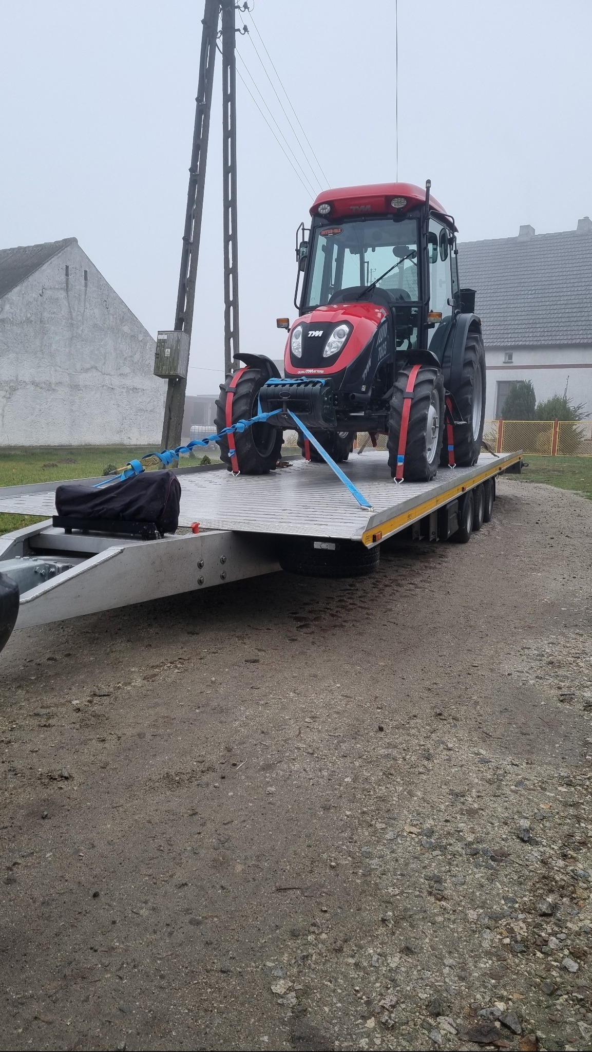 Czerwony traktor TYM zabezpieczony pasami transportowymi na niskopodwoziowej lawecie w pochmurny dzień.