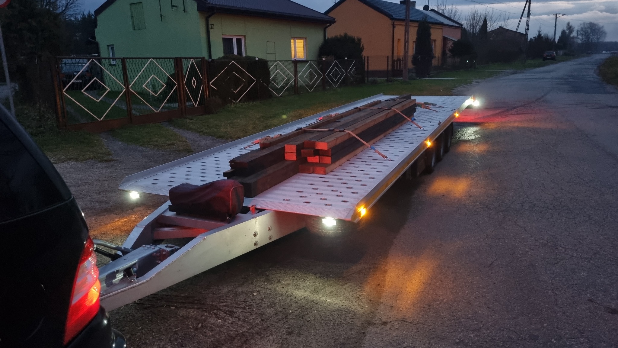 Laweta z drewnianymi belkami zabezpieczonymi pasami transportowymi, zaparkowana na asfaltowej drodze przy zabudowaniach mieszkalnych, oświetlona światłami pozycyjnymi i roboczymi.