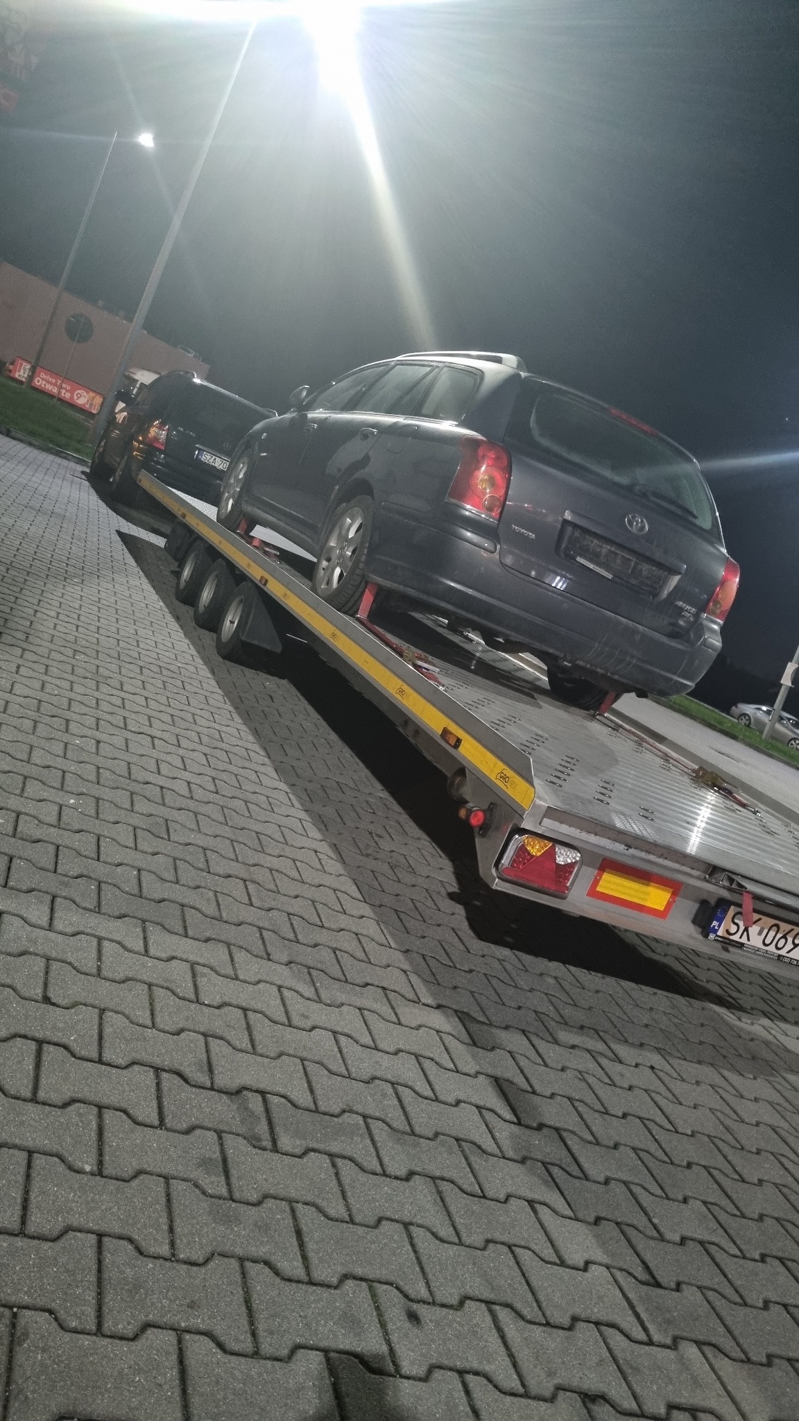 Dwa ciemne samochody, Toyota Avensis i inny sedan, umieszczone na platformie lawety w nocnych warunkach oświetleniowych, widoczne tablice rejestracyjne.