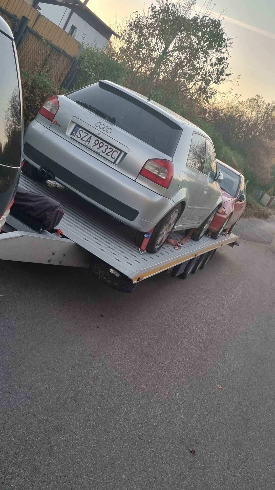 Srebrny samochód Audi A3 z polską tablicą rejestracyjną SZA 9932C, zabezpieczony czerwonymi pasami, transportowany na platformie lawety. W tle widoczny drugi, czerwony samochód oraz zabudowania.