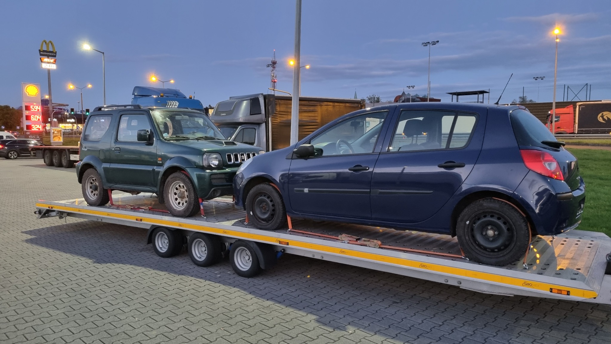 Dwa samochody - zielony Suzuki Jimny i niebieski Renault Clio - zabezpieczone pasami na niskopodwoziowej przyczepie lawety, na tle stacji benzynowej i parkingu dla ciężarówek o zmierzchu.