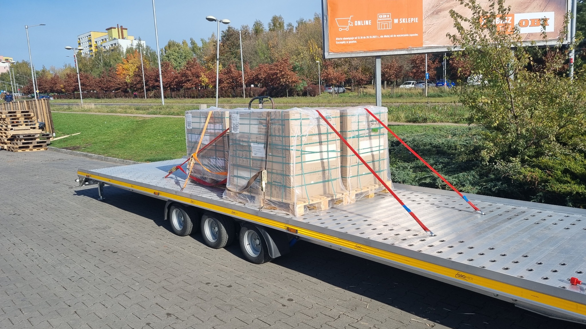 Płaska laweta z trzema paletami zapakowanych towarów zabezpieczonych pasami transportowymi na tle miejskiego krajobrazu z budynkami i billboardem reklamowym.