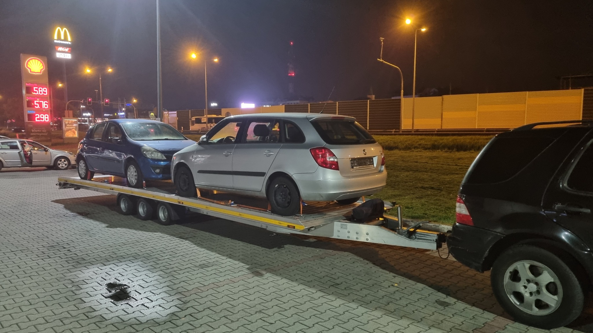 Dwa samochody, srebrny Skoda Fabia i niebieski Renault Clio, zabezpieczone pasami na lawecie, holowane przez czarny samochód osobowy na tle stacji benzynowej i McDonald's o zmierzchu.