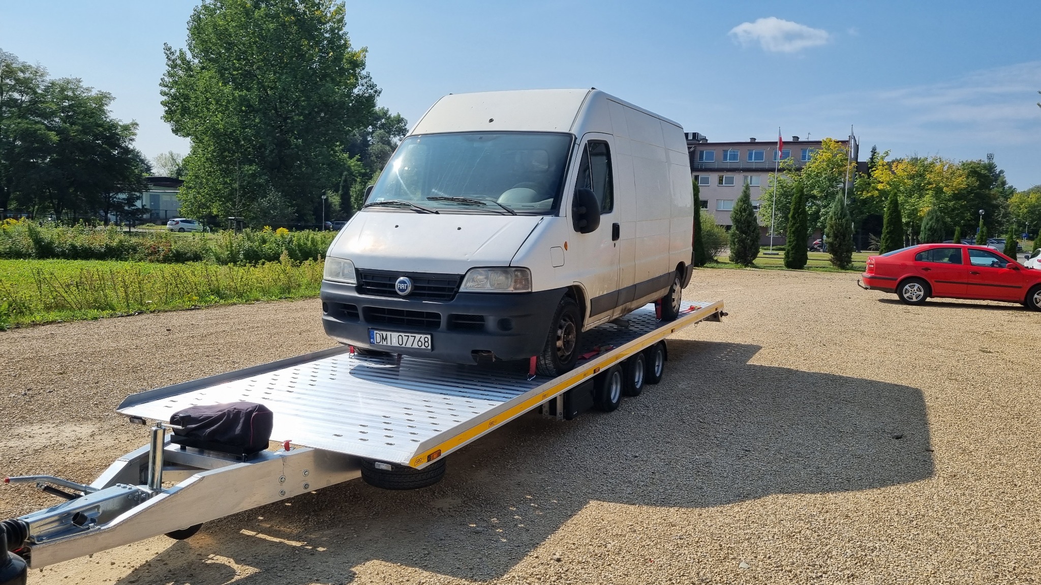 Biały samochód dostawczy marki Fiat Ducato zabezpieczony na srebrnej, trzyosiowej platformie lawety, widok z boku na żwirowym parkingu w słoneczny dzień.