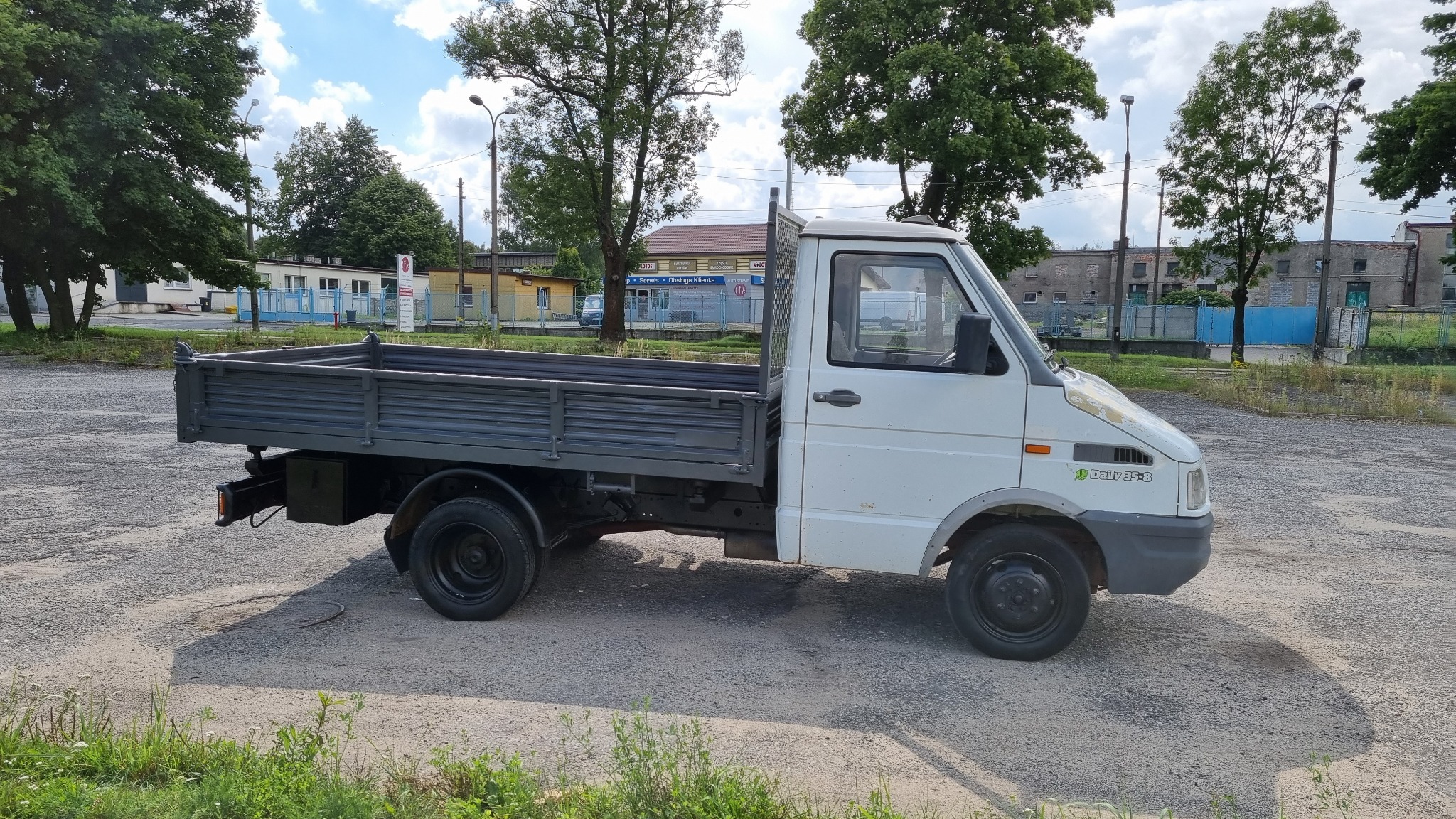 Biała, lekko zniszczona skrzyniowa Iveco Daily 35-8 na szarych, stalowych felgach stoi na żwirowym placu, z widocznymi zabudowaniami w tle.