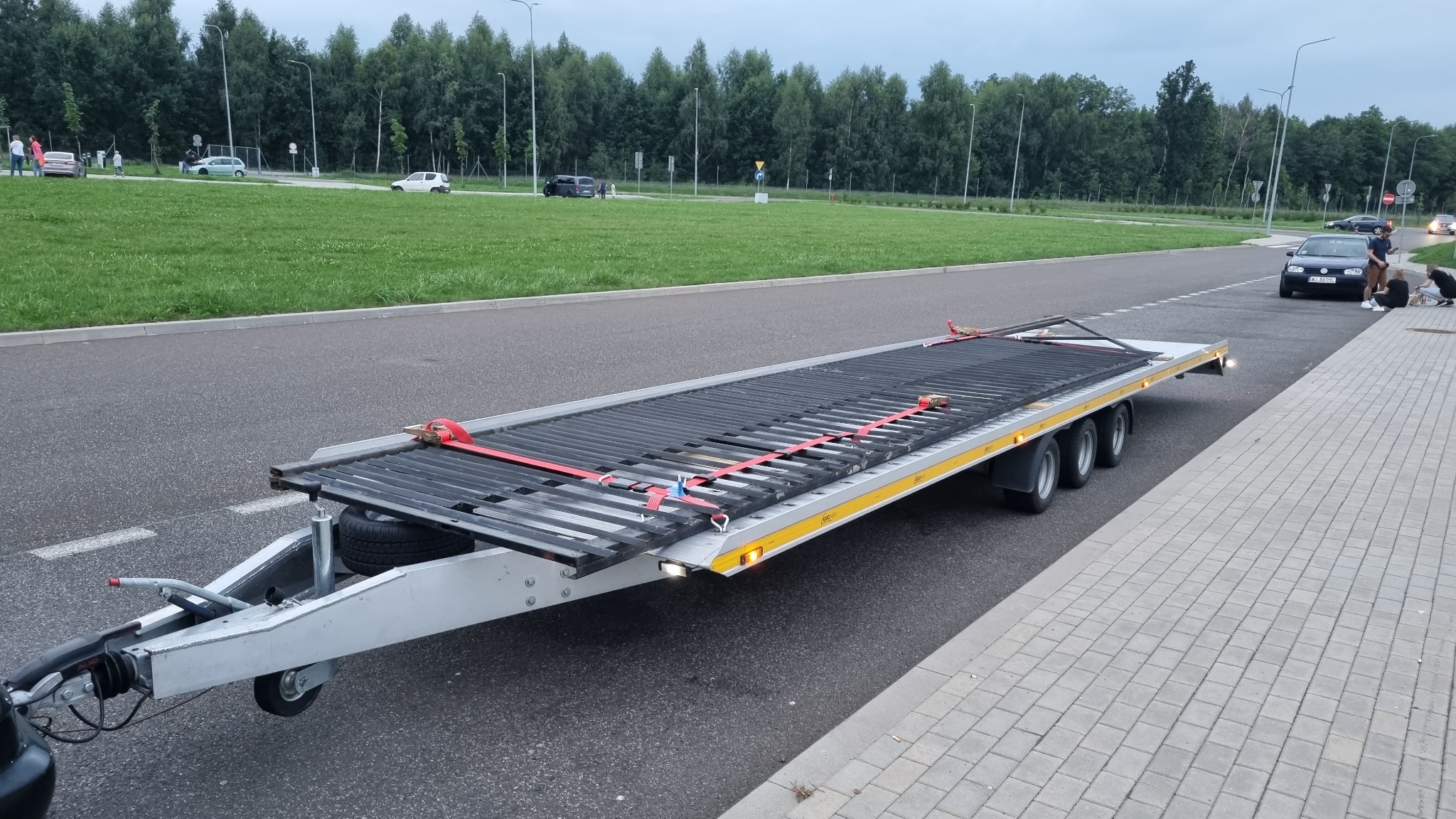 Pusta platforma przyczepy z czarną kratownicą zabezpieczoną czerwonymi pasami, zaparkowana na poboczu drogi, w tle zielony trawnik i drzewa.