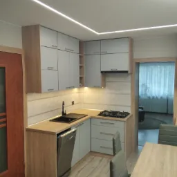 Jasna kuchnia po renowacji z drewnianymi blatami, szarymi szafkami i czarnym zlewem, oświetlenie LED podszafkowe i sufitowe, widoczny fragment stołu i krzeseł.