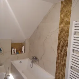 Biała wanna w wyremontowanej łazience na poddaszu ze złotymi mozaikami i grzejnikiem drabinkowym, oświetlenie LED pod wanną.