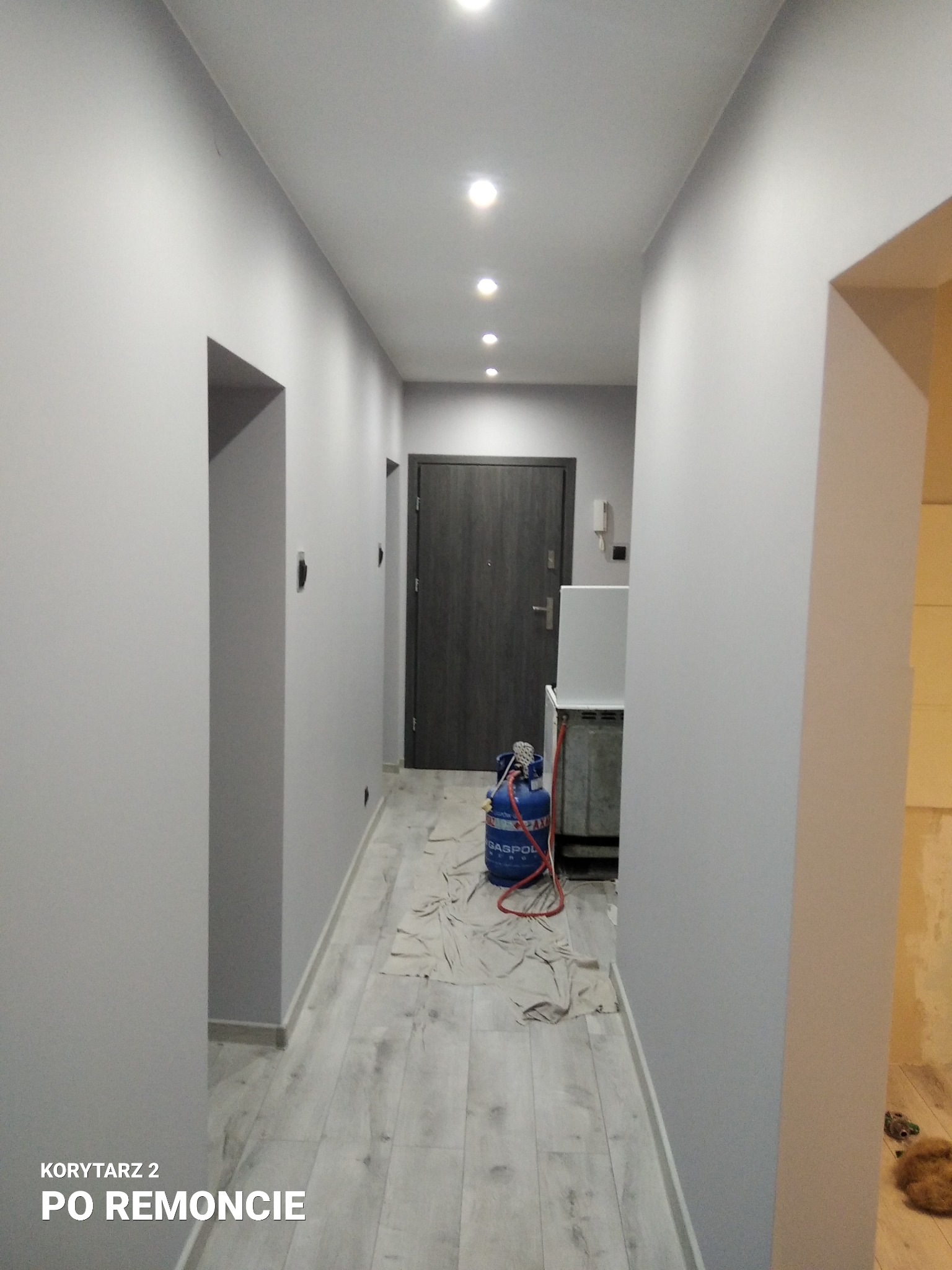 Korytarz w szarościach po remoncie, z widocznymi punktowymi lampami sufitowymi, drzwiami wejściowymi i butlą gazową na podłodze zabezpieczonej folią malarską.