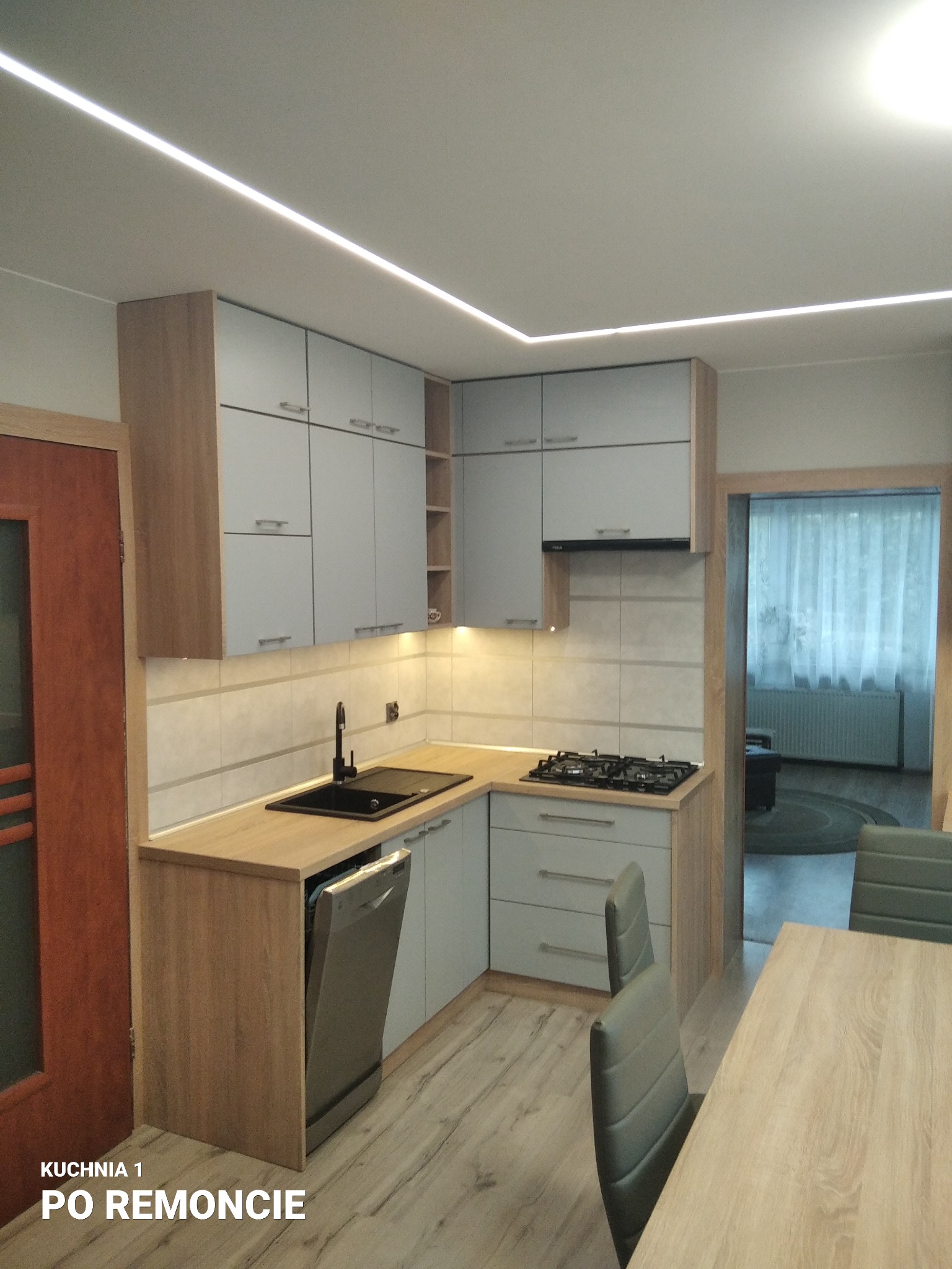 Jasna kuchnia po renowacji z drewnianymi blatami, szarymi szafkami i czarnym zlewem, oświetlenie LED podszafkowe i sufitowe, widoczny fragment stołu i krzeseł.