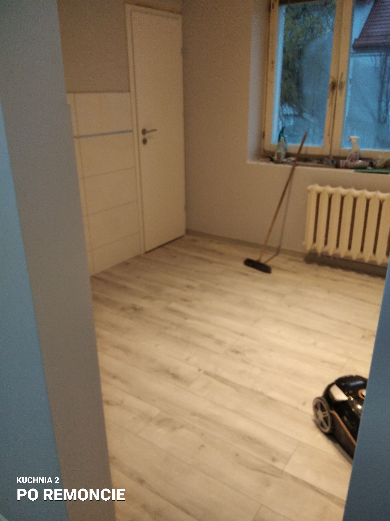 Wykończona kuchnia z panelami podłogowymi w odcieniach beżu i szarości, widoczny odkurzacz automatyczny, grzejnik i okno z widokiem na zewnątrz, ściany pomalowane na szaro.