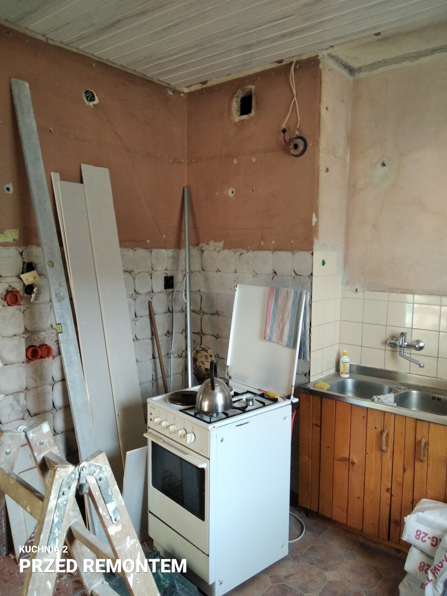 Kuchnia w trakcie przygotowań do remontu: widoczne odsłonięte ściany, stara zabudowa kuchenna, drabina malarska oraz materiały budowlane.