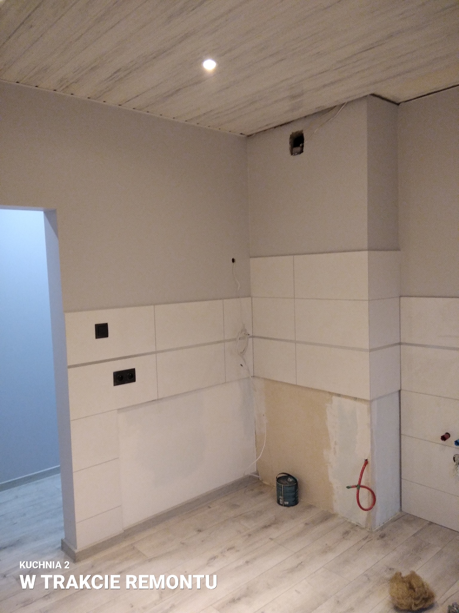 Kuchnia w trakcie remontu: częściowo położone białe płytki na ścianie, widoczne gniazdka elektryczne, fragment instalacji hydraulicznej, puszka farby na podłodze z paneli imitujących drewno, sufit...