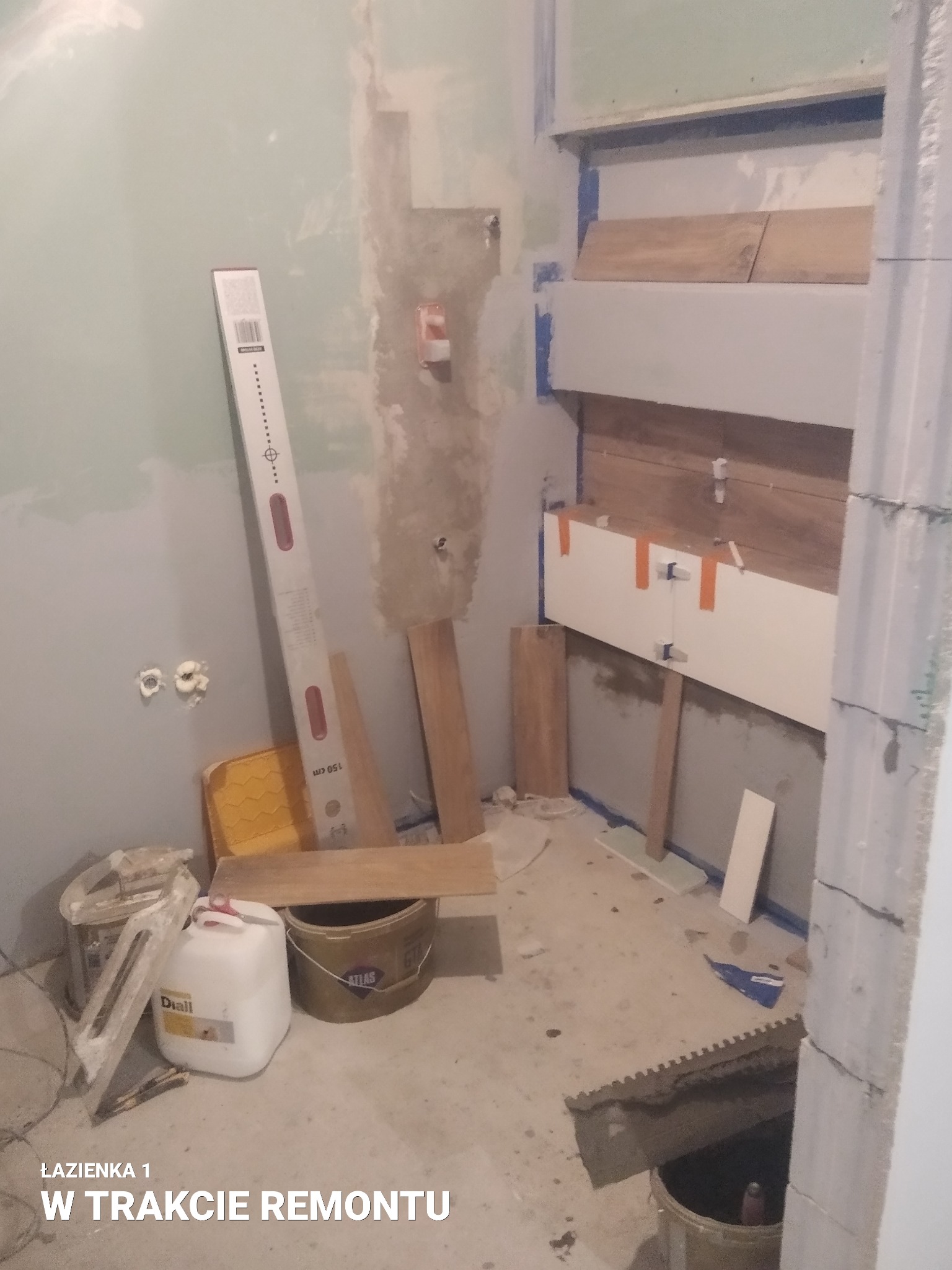 Łazienka w trakcie prac: montaż płytek imitujących drewno na ścianie, widoczne poziomica, wiadro z klejem Atlas, pojemnik Diall, fragment ściany z płyt karton-gips i instalacją elektryczną.