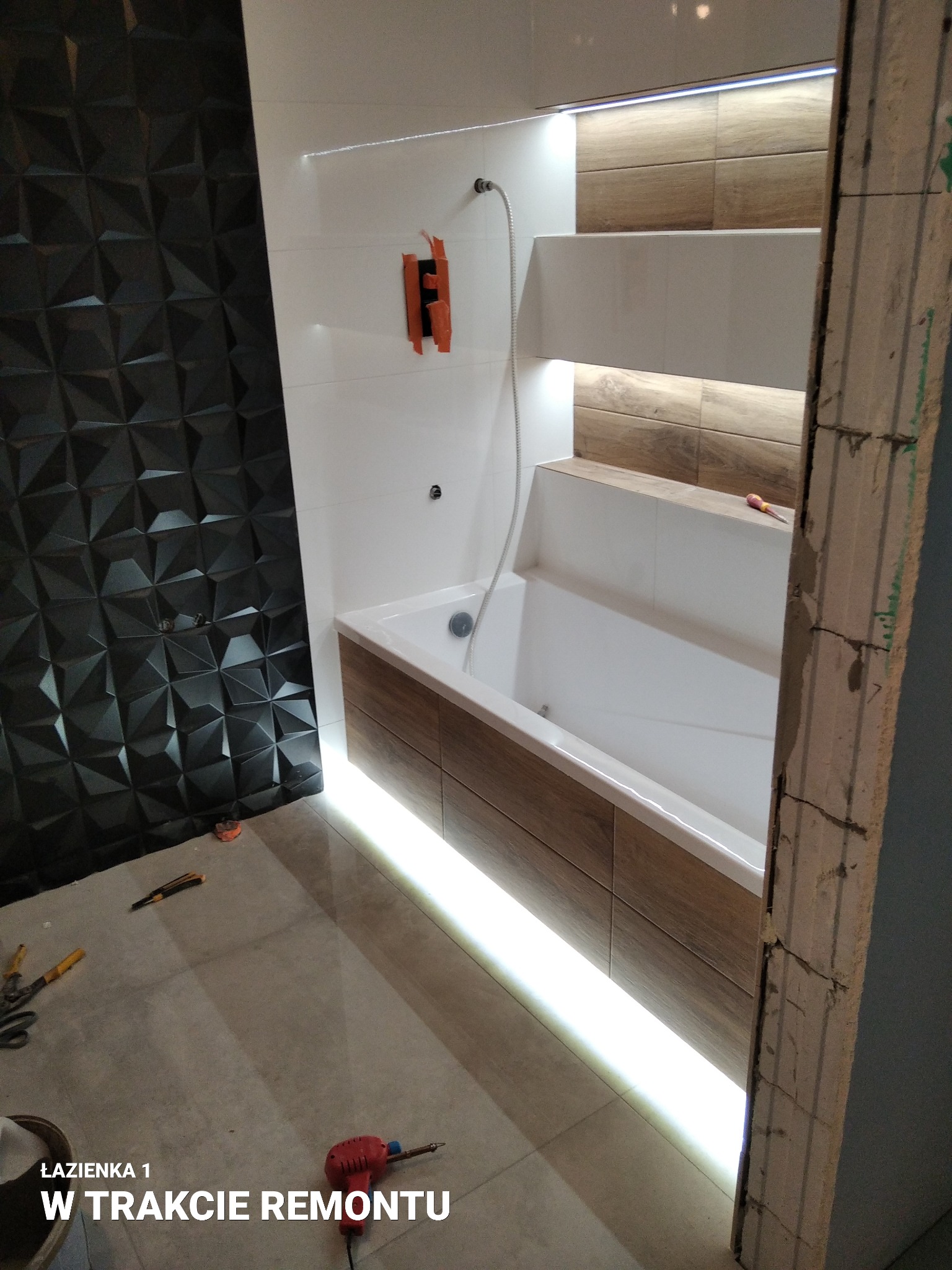 Łazienka w trakcie prac: czarna trójwymiarowa ściana, biała wanna z drewnopodobną obudową podświetloną LED, półki z podświetleniem, widoczne narzędzia na podłodze i surowy fragment ściany.