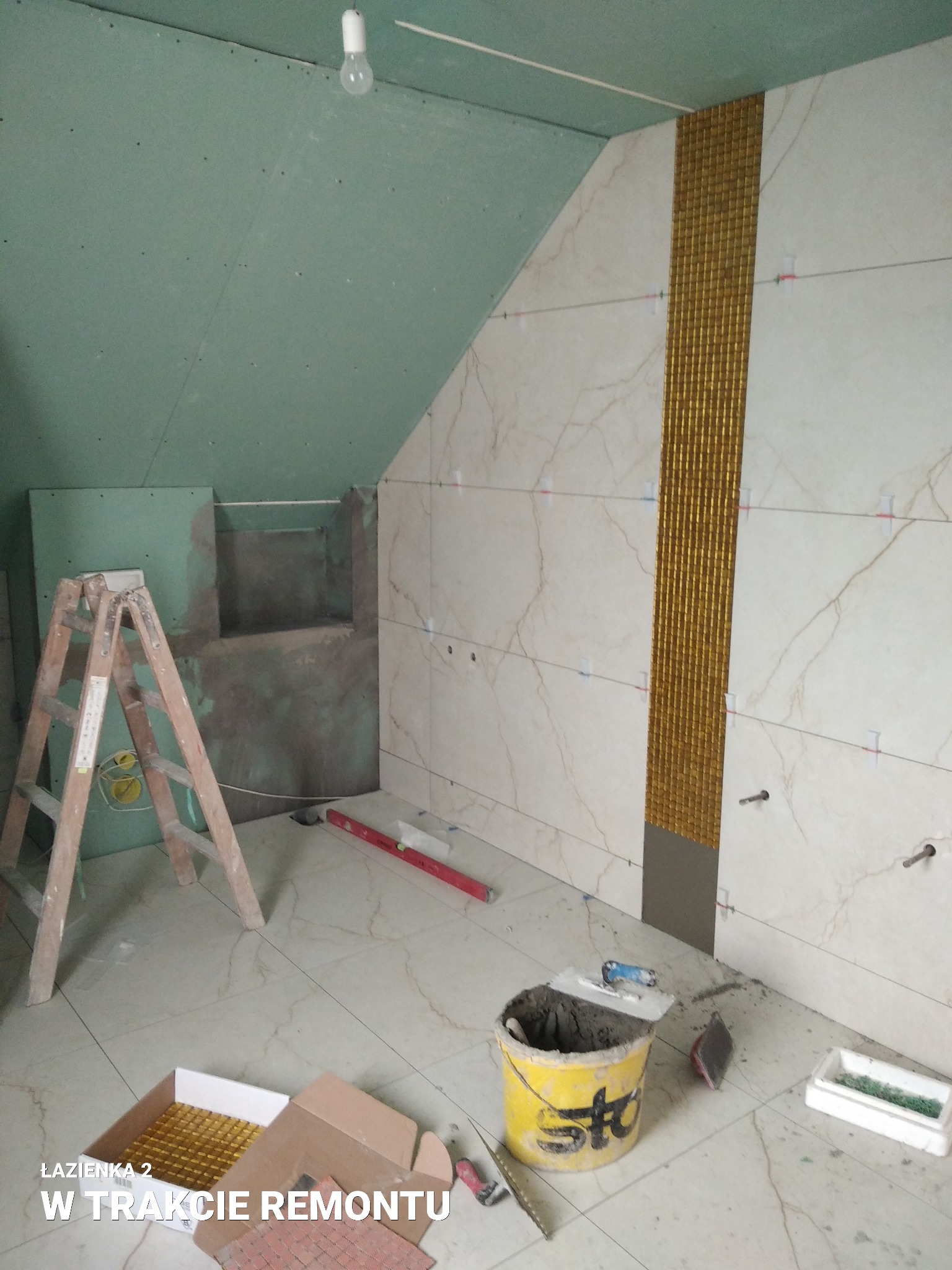 Łazienka na poddaszu w trakcie układania płytek: ściany wyłożone marmurkowymi płytkami z pionowym pasem złotej mozaiki, widoczny drabina, poziomica, wiadro z zaprawą i pudełko z mozaiką.