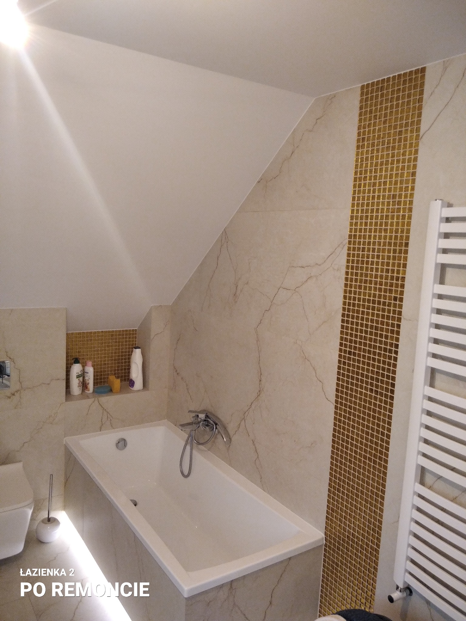 Biała wanna w wyremontowanej łazience na poddaszu ze złotymi mozaikami i grzejnikiem drabinkowym, oświetlenie LED pod wanną.