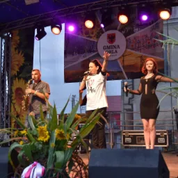 Występ na scenie: wokalistka w białej koszulce z napisem, saksofonista i tancerka w czarnej sukience na tle baneru z herbem Gminy Orla.