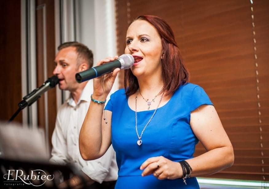 Wokalistka w niebieskiej sukience śpiewa do mikrofonu, drugi wokalista w białej koszuli w tle, brązowe żaluzje w tle