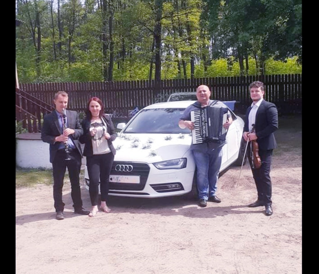 Czwórka muzyków z klarnetem, akordeonem i skrzypcami pozuje przed udekorowanym białym Audi A4 w plenerze.