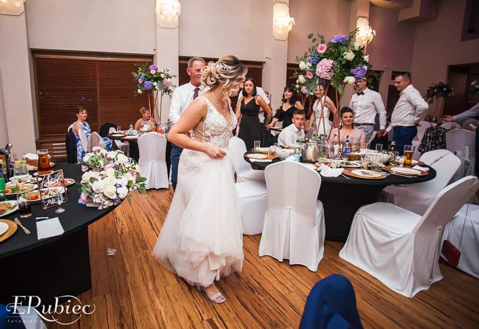 Panna młoda w białej sukni przechadza się po sali weselnej, w tle goście siedzący przy okrągłych stołach z czarnymi obrusami i kwiatowymi dekoracjami.