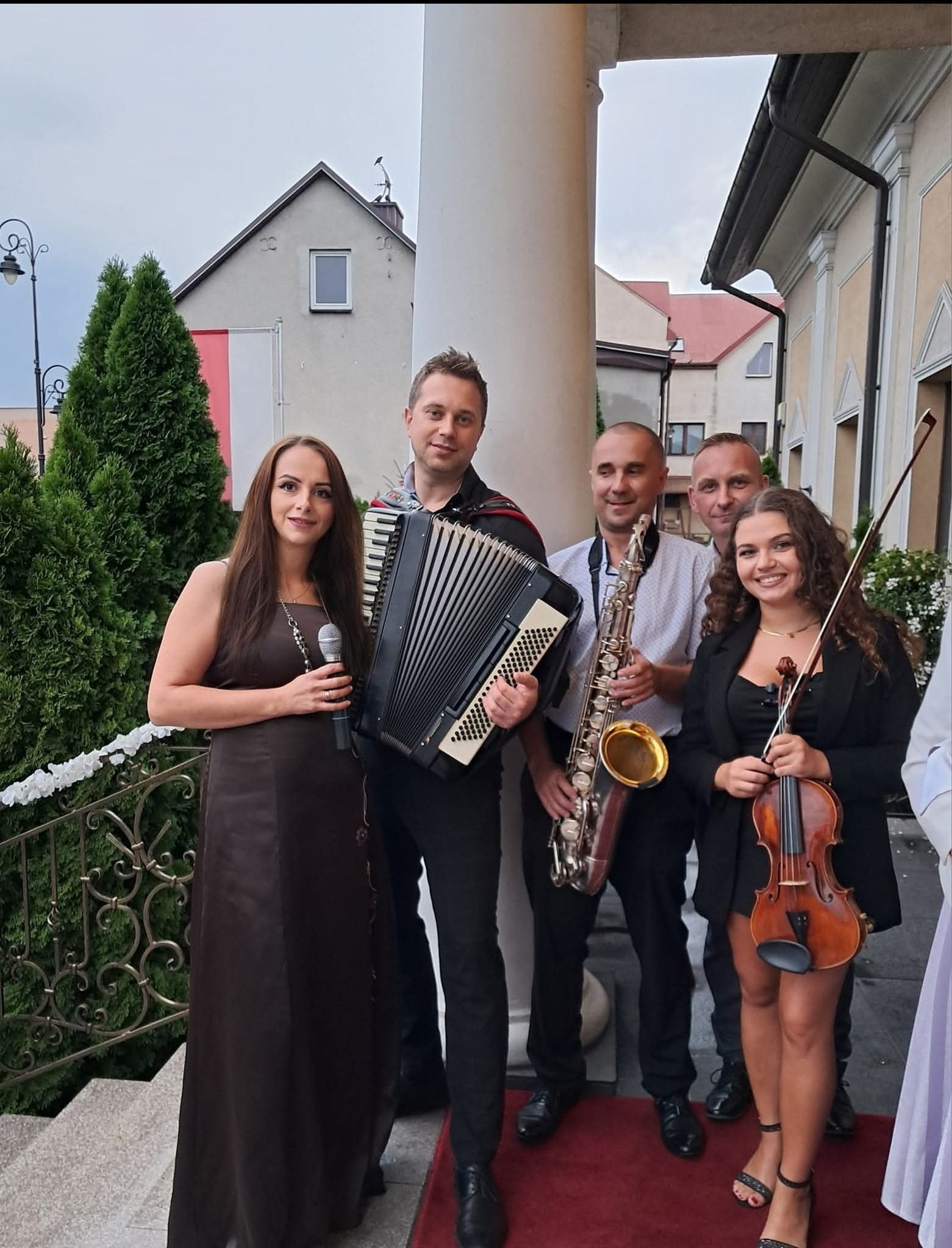 Zespół muzyczny: wokalistka z mikrofonem, akordeonista, saksofonista i skrzypaczka na schodach przed budynkiem. Uśmiechnięci muzycy w strojach scenicznych.