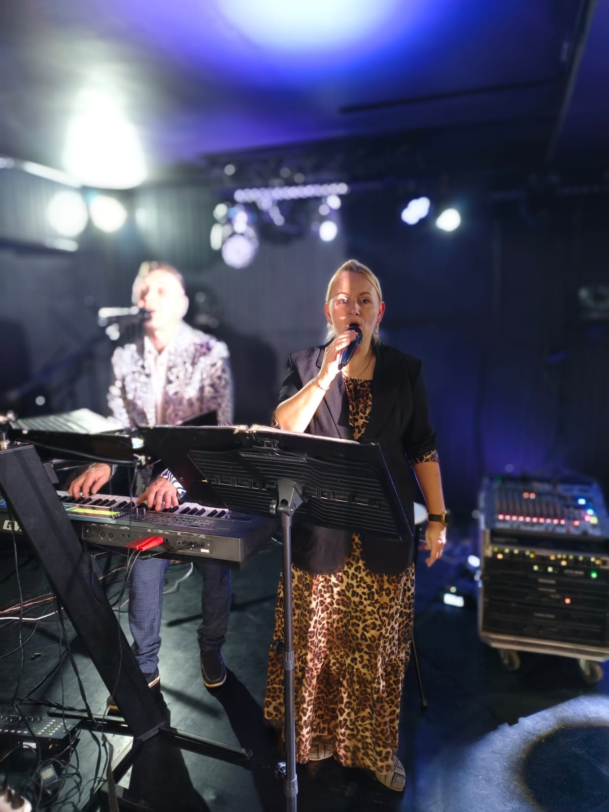Wokalistka w sukience w panterkę śpiewa do mikrofonu, obok klawiszowiec w kwiecistej marynarce. Scena oświetlona niebieskim światłem, widoczny mikser dźwięku.