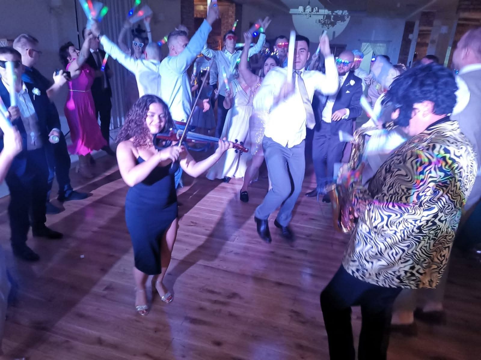 Skoczna zabawa z muzyką na żywo: skrzypaczka w czarnej sukience i saksofonista w wzorzystej marynarce, rozbawiony tłum z ledowymi pałeczkami w tle.