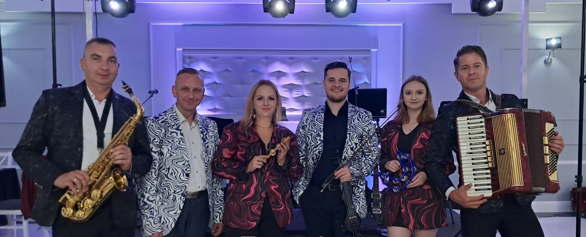 Zespół muzyczny w kolorowych marynarkach prezentuje instrumenty: saksofon, akordeon, skrzypce elektryczne i tamburyn. Scena oświetlona reflektorami.