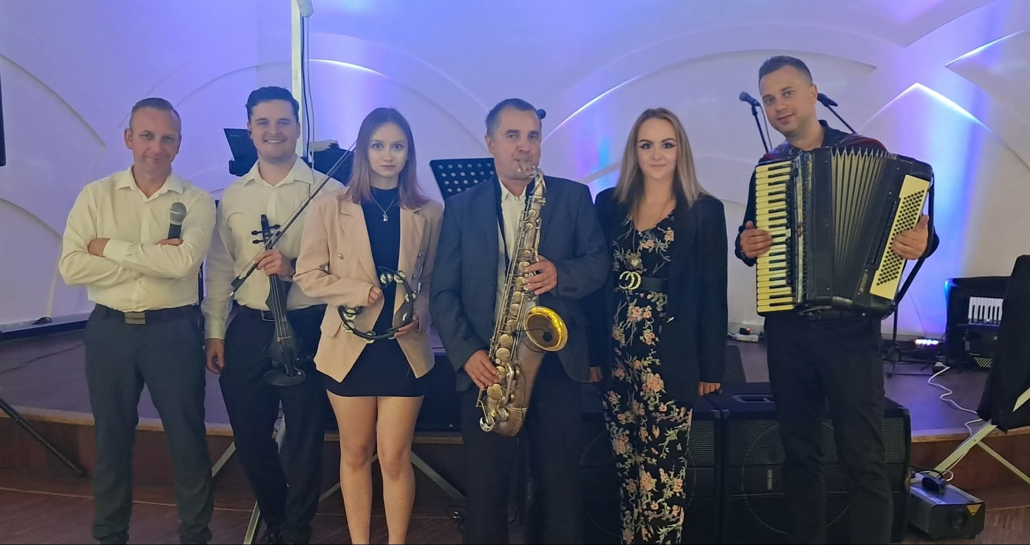 Zespół muzyczny w składzie sześcioosobowym, z saksofonem, akordeonem, skrzypcami i tamburynem, pozuje na scenie z niebieskim oświetleniem w tle.
