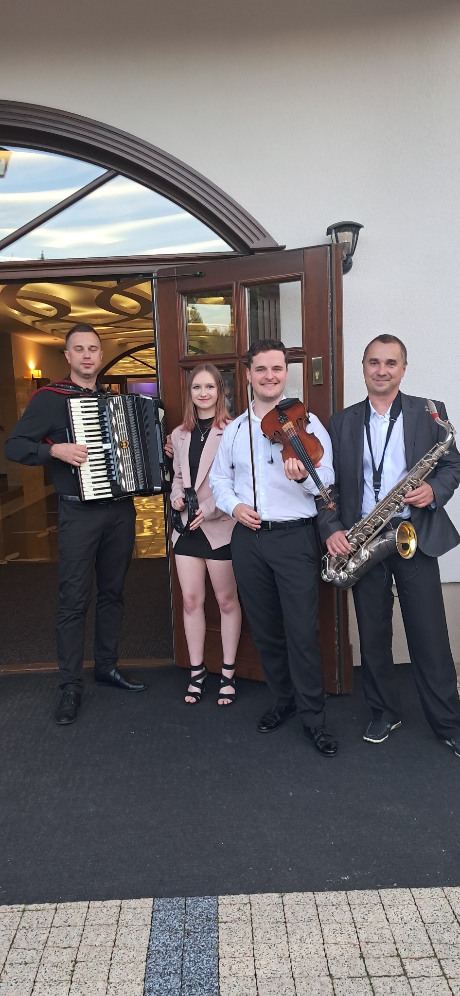 Zespół muzyczny: akordeonista, skrzypek, saksofonista i wokalistka pozują przed wejściem do budynku z dekoracyjnymi drzwiami.